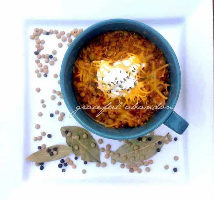 bowl of lentil chili on white plate with bay leaves, lentils, and peppercorns on white plate for visual interest